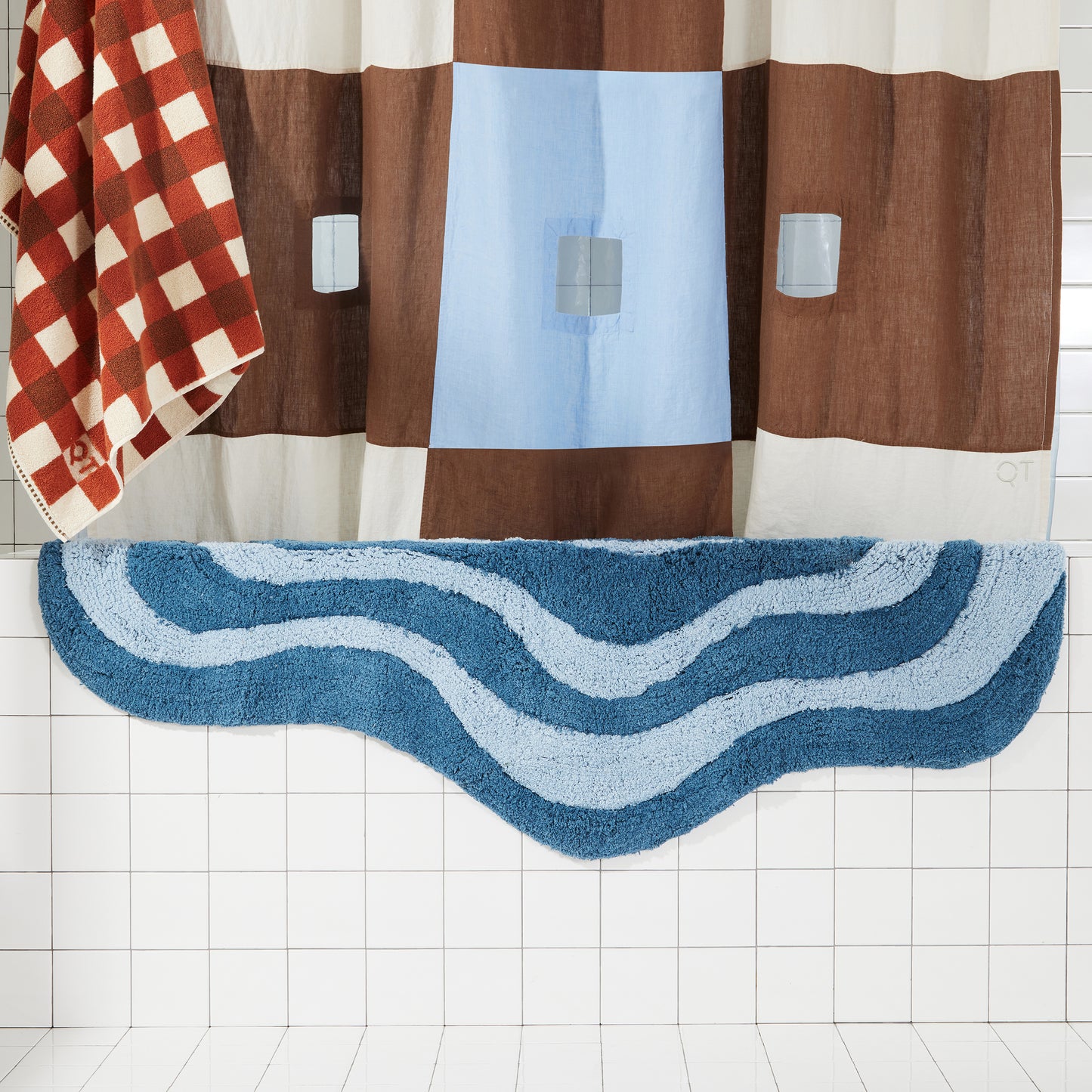 dark and light blue wavy bath rug draped over white tiled bath tub next to brown and blue shower curtain and clay and brown bath sheet. 