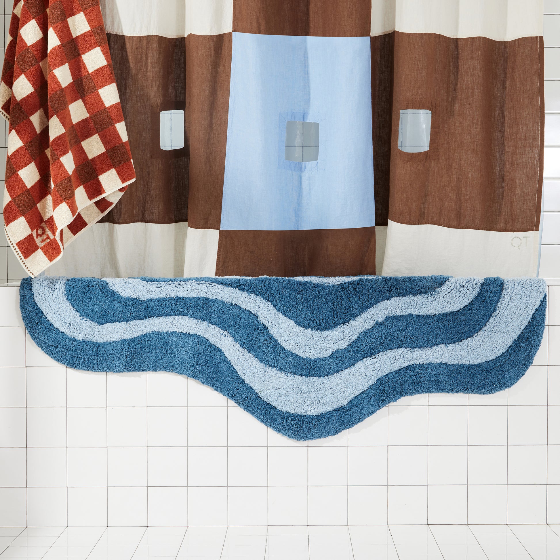 dark and light blue wavy bath rug draped over white tiled bath tub next to brown and blue shower curtain and clay and brown bath sheet. 