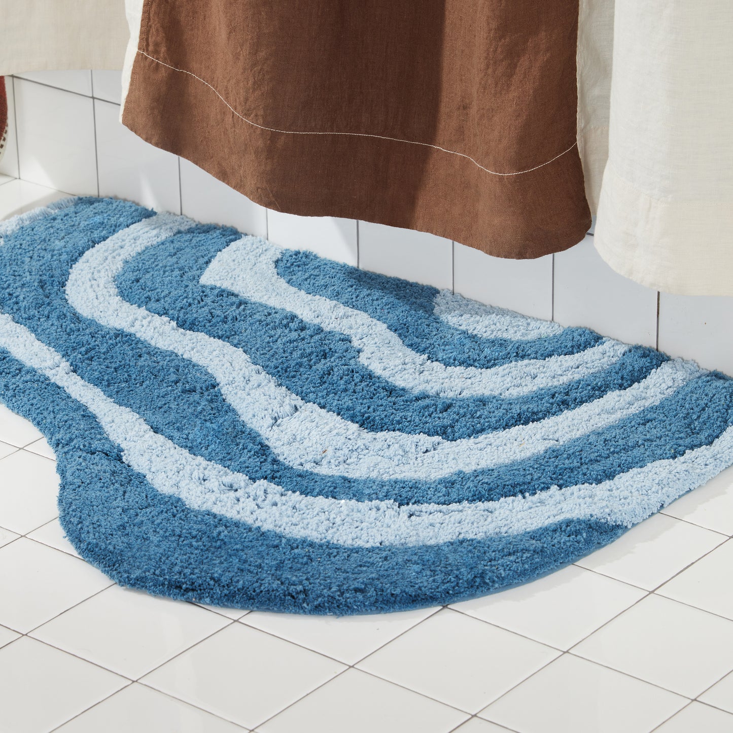 dark and light blue wavy bath rug on white tiled floor next to brown and cream shower curtain. 