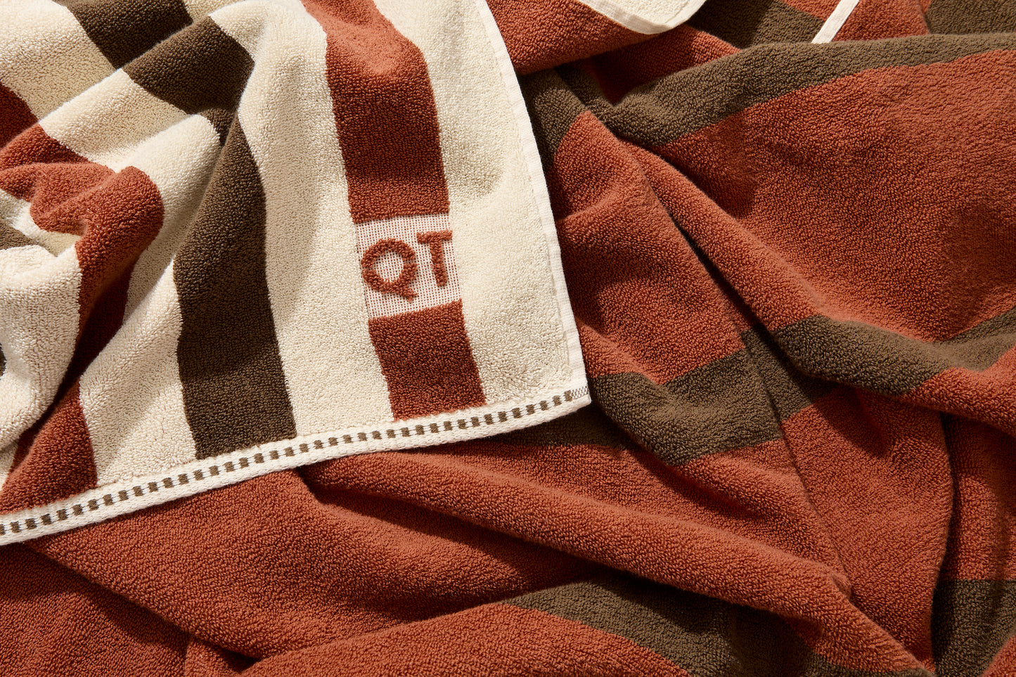 Corner detail of catalina bath sheet in chocolate with embossed QT logo on one side and color to color stripes on other side. 