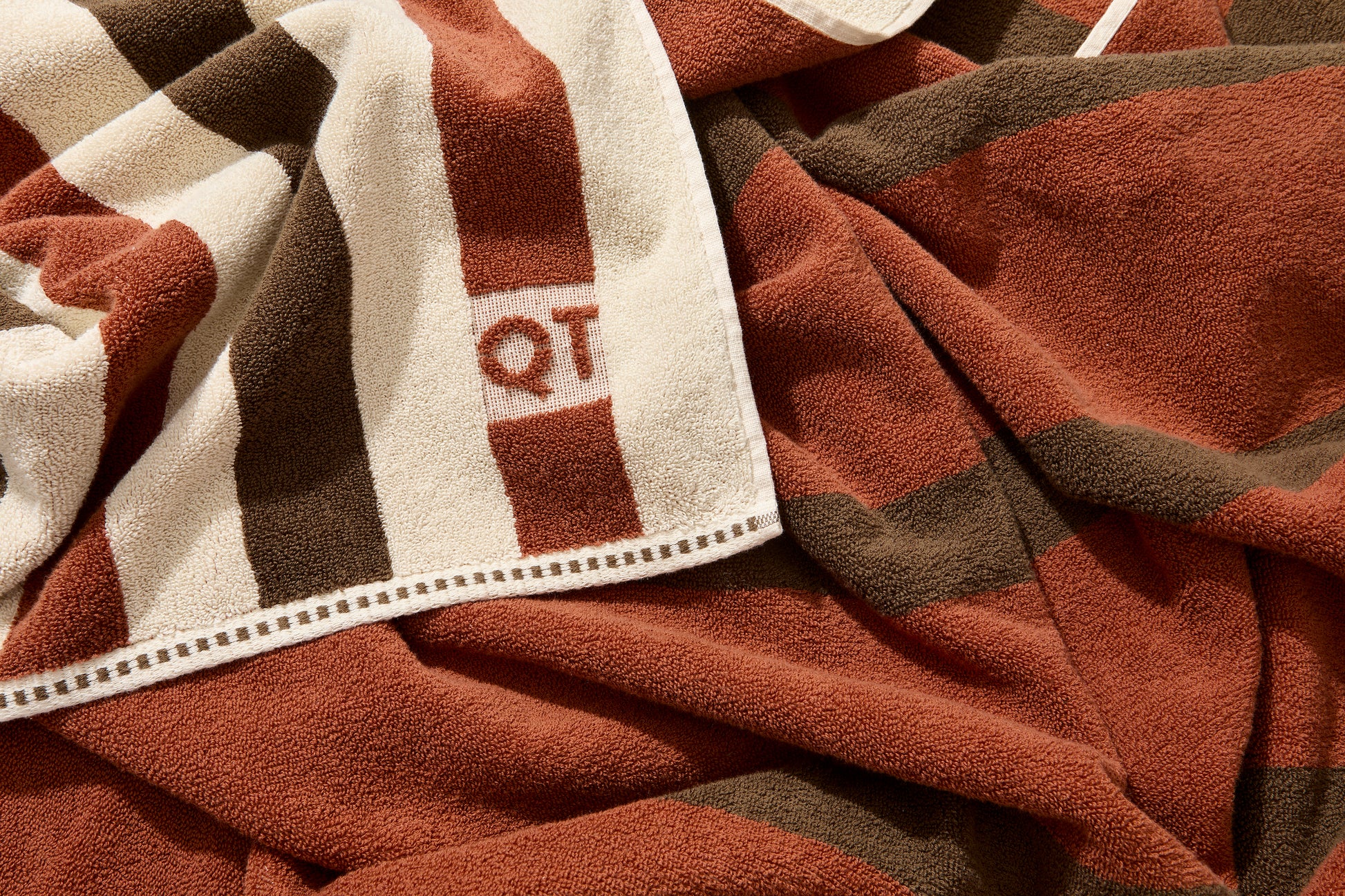 Corner detail of catalina bath sheet in chocolate with embossed QT logo on one side and color to color stripes on other side. 